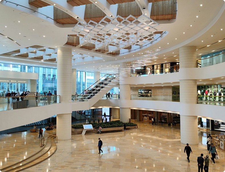 Modern commercial building interior with spacious lobby, elegant lighting fixtures, and glass walls, showcasing a busy environment with people and retail spaces.