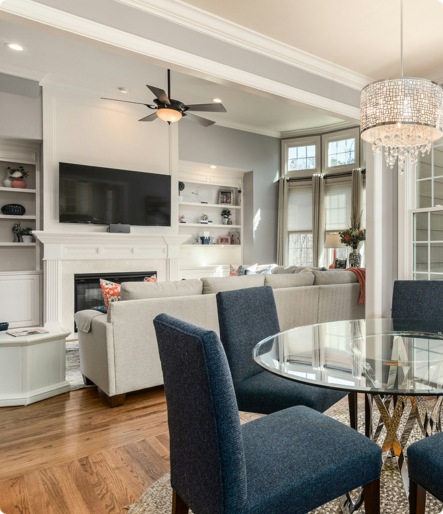 Modern living room with a light-colored sofa, glass dining table, and blue upholstered chairs, featuring a ceiling fan and a chandelier, designed to complement Florida's climate and aesthetic preferences.