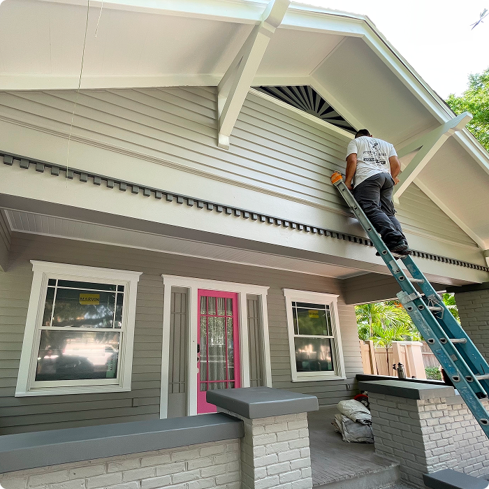 Worker applying exterior paint on a house in Florida, using a ladder, showcasing meticulous surface preparation and detail-oriented application for enhanced weather protection.
