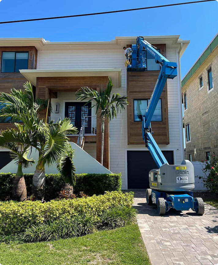 Worker using a lift for exterior painting on a modern Florida home, featuring tropical landscaping and contrasting wooden accents.