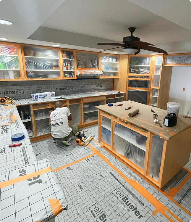 Painter preparing surfaces in a kitchen, with cabinets covered in plastic sheeting, using tools on a protected floor, emphasizing meticulous surface preparation for a painting project.