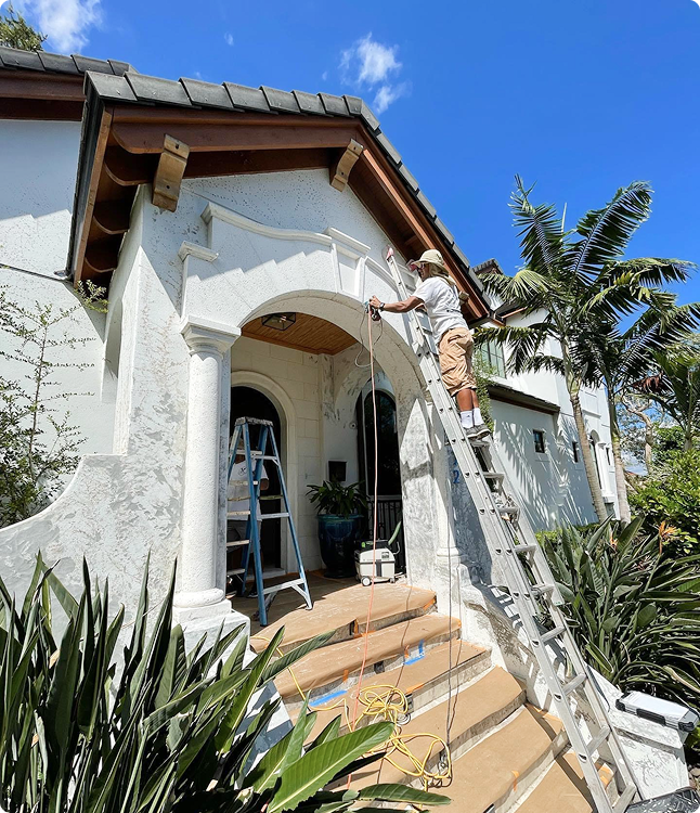 Professional painter on a ladder applying paint to the exterior of a Florida home, showcasing meticulous preparation and attention to detail in a sunny environment with tropical plants.
