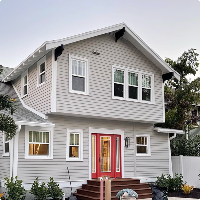 Freshly painted two-story house with gray siding and red front door, showcasing meticulous exterior preparation for Florida's climate, surrounded by greenery and construction materials.