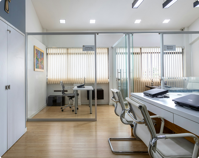 Modern office interior with glass partitions, featuring a desk and chair in a private workspace, and a collaborative area with laptops, emphasizing a professional environment for commercial operations.