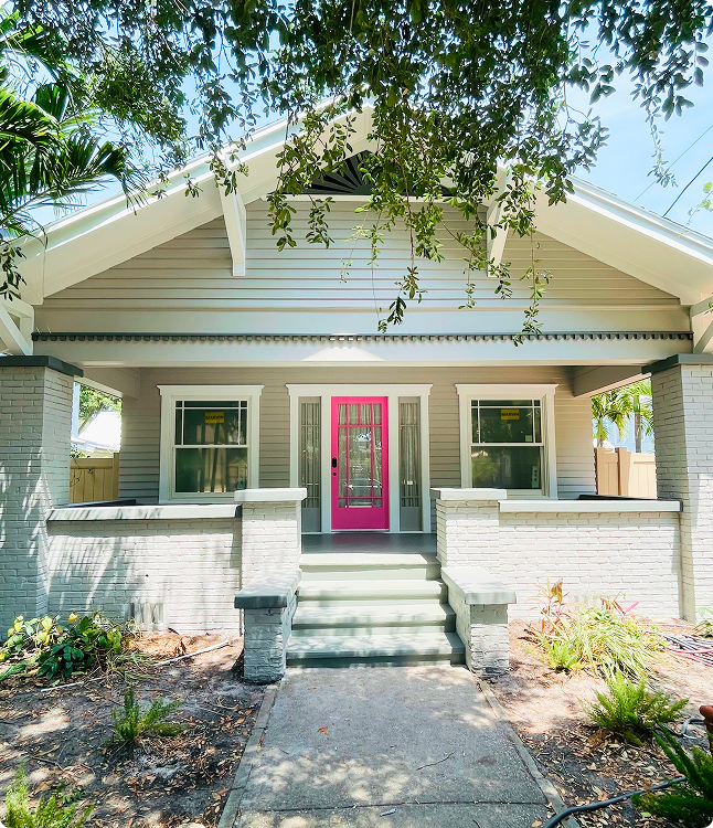 Exterior view of a Florida home featuring a vibrant pink front door, gray siding, and stone accents, surrounded by lush greenery, showcasing the quality and detail of Attention to Detail Painting's work.