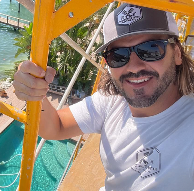 Man smiling and holding a scaffold railing, wearing sunglasses and a "Attention to Detail" shirt, with a swimming pool and palm trees in the background, showcasing the company's commitment to quality work in a vibrant setting.