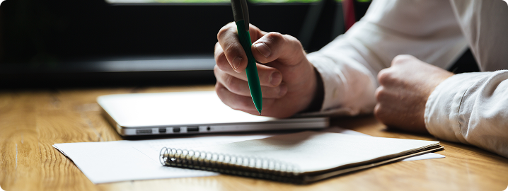 Person writing in a notebook with a green pen, laptop and blank papers on a wooden table, emphasizing property maintenance and painting checklist tasks for homeowners.