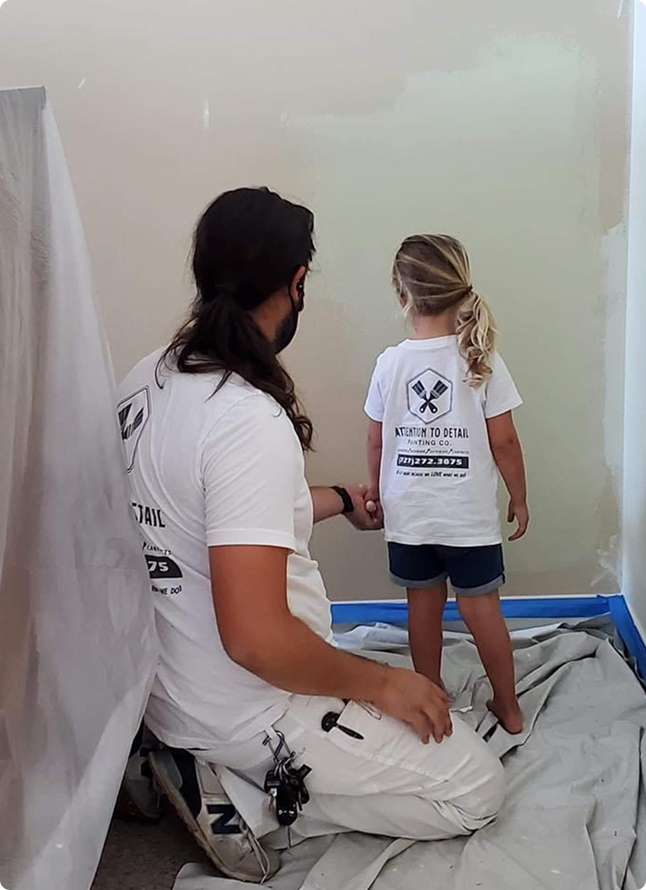 Adult painter and child in matching "Attention to Detail Painting Co." shirts, preparing a wall for painting in a coastal home setting.
