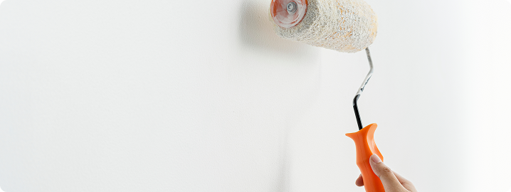Hand holding a paint roller applying paint to a white wall, illustrating surface preparation in European painting techniques.